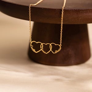 May include: A gold-toned necklace featuring three interlocking heart-shaped pendants. Each heart is inscribed with a name in cursive script: Eartha, Clara, and Denise. The pendant is suspended from a delicate gold-toned chain link necklace.