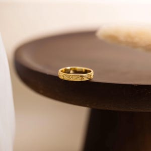 May include: A gold band ring featuring a continuous engraved pattern of swirling wave-like lines and small circular dots. The metal surface has a polished finish, and the edges of the ring are detailed with a fine beaded texture.
