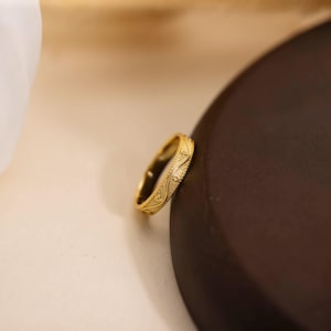 May include: A gold ring with an intricate, repeating pattern. The ring is set against a neutral background, highlighting its detailed design. The ring's surface reflects light, showcasing its polished finish. The ring appears to be a band, suitable for everyday wear.