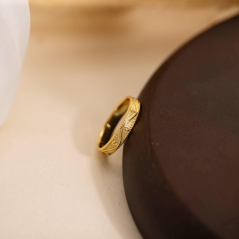 May include: A gold ring with an intricate, repeating pattern. The ring is set against a neutral background, highlighting its detailed design. The ring's surface reflects light, showcasing its polished finish. The ring appears to be a band, suitable for everyday wear.