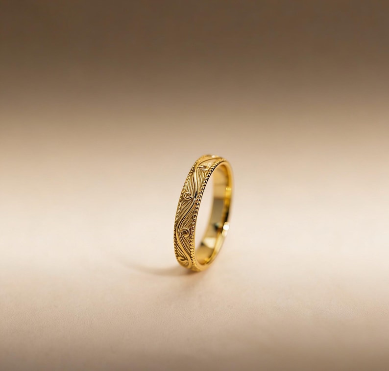 May include: A gold ring featuring a polished finish and a central band decorated with an intricate, repeating engraved swirl pattern, bordered on both sides by a delicate beaded edge detail that runs along the entire circumference.