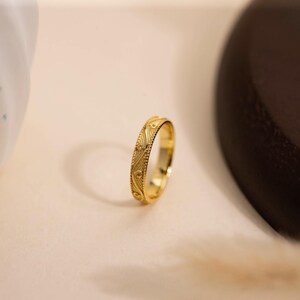 May include: A gold ring with an intricate, repeating pattern. The ring is a bright, shiny gold color and appears to be made of metal. The design features a series of curved lines and small dots. The ring is set against a neutral background.