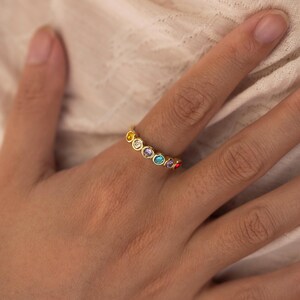 Può includere: Un anello d'oro con una serie di pietre preziose colorate. L'anello ha una fascia liscia e lucida ed è adornato con pietre rotonde di vari colori, tra cui giallo, bianco, blu e rosso. Le pietre preziose sono poste vicine, creando un design vibrante e accattivante.