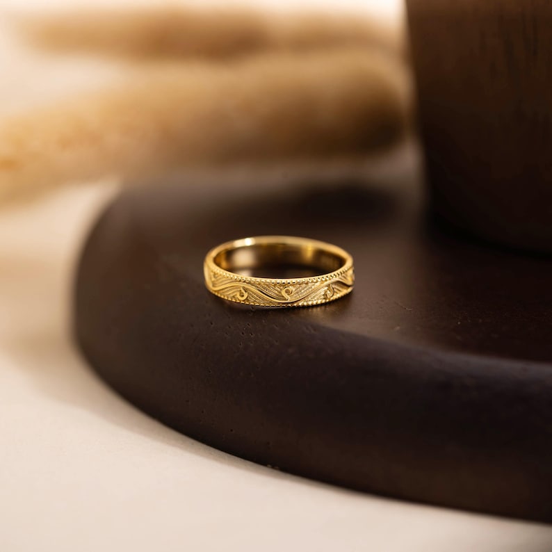 May include: A gold-colored ring with an intricate wave and dot pattern. The ring is set on a dark brown surface, with a blurred background. The ring's design features a repeating pattern of curved lines and small dots.