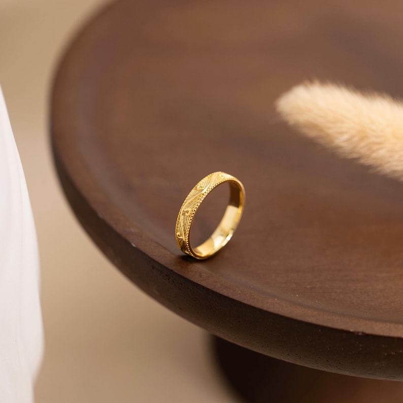 May include: A gold ring with an intricate pattern of small dots and lines. The ring is set on a dark brown wooden surface. The ring is a band style, with a continuous design around the circumference.