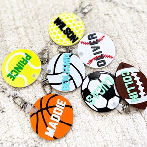 May include: A collection of sports-themed keychains. The keychains feature designs of a golf ball, baseball, tennis ball, volleyball, basketball, soccer ball, and football. Each keychain has a name printed on it in a contrasting color.
