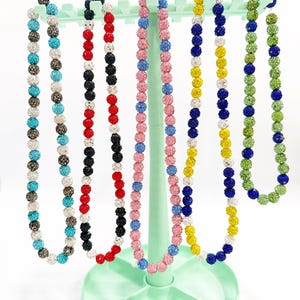 May include: A display of beaded necklaces in various colors, including turquoise, black, red, pink, yellow, blue, and green. The necklaces are displayed on a light green jewelry stand. Each necklace features a series of small, round beads.