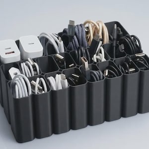 Peut inclure: Un organisateur de câbles en plastique noir doté de multiples compartiments rectangulaires contenant divers accessoires électroniques, notamment des adaptateurs secteur blancs, des câbles de charge blancs enroulés et des cordons USB noirs.