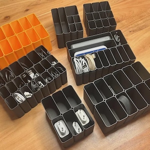 May include: A collection of modular plastic drawer organizers in bright orange and dark gray. These rectangular grid containers feature multiple compartments of varying sizes, some holding coiled white charging cables, power adapters, and small electronic devices.