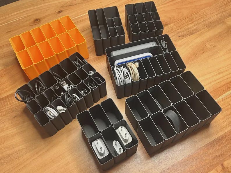 May include: A collection of modular plastic drawer organizers in bright orange and dark gray. These rectangular grid containers feature multiple compartments of varying sizes, some holding coiled white charging cables, power adapters, and small electronic devices.