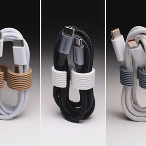 May include: Three coiled USB-C charging cables secured with ribbed silicone cable organizers. The first is a white cable with a tan organizer, the second is a black UGREEN cable with a white organizer, and the third is a white cable with a gray organizer.