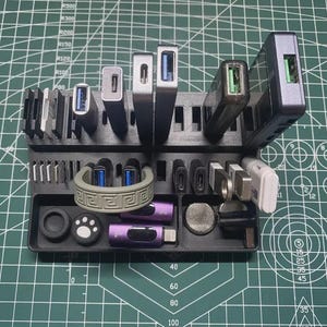 May include: A black 3D-printed plastic organizer stand featuring multiple slots for various USB flash drives, adapters, and small electronic accessories. The stand holds several metallic and plastic devices, including purple, silver, and white components.