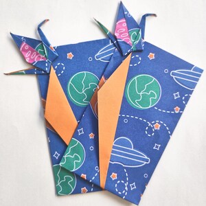 May include: Two origami paper cranes atop folded blue paper with a space-themed pattern. The paper features planets, stars, and UFOs. Orange paper accents the folds, creating a striking contrast. The cranes are blue with pink accents.