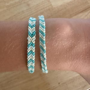 May include: Two handmade woven bracelets made of light blue and cream-coloured thread. One bracelet features a chevron pattern, while the other displays a diagonal striped pattern. Both bracelets are thin and constructed from knotted embroidery floss.