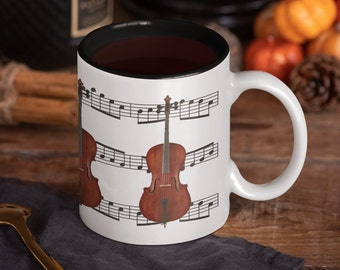 May include: White ceramic mug with a black rim. The mug features a design of three cellos on a background of musical notes. The cellos are brown and have a natural wood finish.