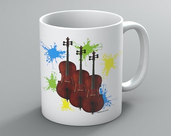 May include: White ceramic mug featuring a design of three brown cellos with black fingerboards and colorful paint splatters in blue, green, and yellow. The mug has a white handle and a glossy finish.