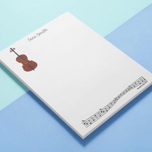 May include: White notepad with a watercolor illustration of a violin and musical notes. The name "Jane Smith" is written in cursive at the top. The musical notes are printed in black ink. The notepad is on a blue and teal background.