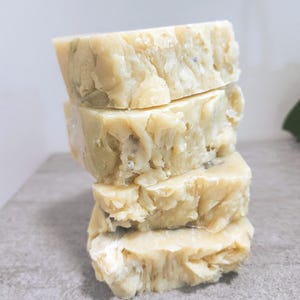May include: Four handmade soap bars stacked on top of each other. The soaps are a pale beige colour with visible flecks of green and purple. The edges of the bars have a rough, textured appearance, suggesting a natural, artisanal quality. The soaps are on a grey surface.
