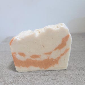 May include: A rectangular bar of handmade soap with a light beige base and swirling orange-red patterns. The top edge has a rough, uneven texture. The soap is sitting on a gray surface.
