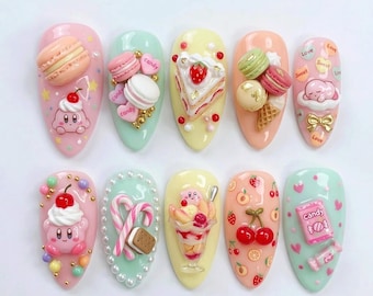 May include: A collection of ten artificial nails, each adorned with miniature, colorful dessert-themed decorations. Designs include macarons, cake slices, ice cream sundaes, and cartoon characters, with accents of candy hearts and cherries.