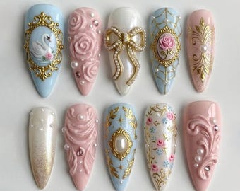 May include: A set of ten almond-shaped press-on nails featuring intricate 3D designs. Embellishments include gold filigree frames, sculpted pink roses, pearl-studded bows, a swan illustration, floral patterns, and gold leaf accents on pastel bases.