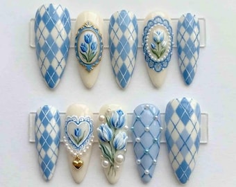 Può includere: Un set di dieci unghie finte a forma di mandorla. I disegni includono motivi a rombi blu e bianchi, motivi floreali e dettagli di perle. Alcune unghie presentano tulipani 3D e ciondoli a forma di cuore, in tonalità pastello.