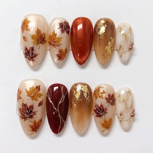 May include: A set of ten almond-shaped press-on nails featuring autumn-themed designs. The nails display cream bases with painted maple leaves in orange and burgundy, solid metallic copper, brown with gold leaf flakes, and white marble patterns.