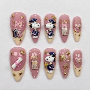 May include: A set of ten artificial nails with a graduation theme. The nails are primarily pink with white tips and feature Snoopy in a graduation cap, books, scrolls, and the words "GRAD" and "CLASS OF". Gold stars and hearts are also included.