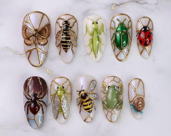 Puede incluir: Una colección de diez uñas artificiales, cada una adornada con un diseño de insecto diferente. Los insectos incluyen una polilla, una abeja, una mantis religiosa, un escarabajo, una mariquita, una araña, una cigarra, una abeja, una rana y un caracol. Cada uña tiene un contorno dorado.