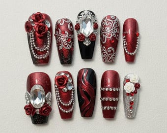 May include: A set of ten artificial nails in various designs. The nails feature a combination of red, black, and silver colors, with embellishments such as rhinestones, pearls, and floral accents. Some nails have intricate patterns and textures.