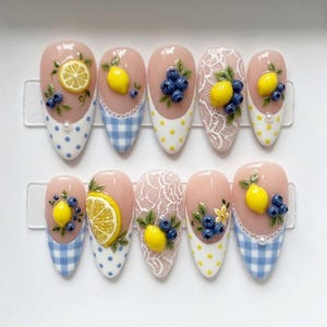May include: A set of ten almond-shaped artificial nails. Each nail features a light pink base with a decorative design of lemons, blueberries, and lace. Some nails have a blue and white gingham or polka dot pattern at the tips.