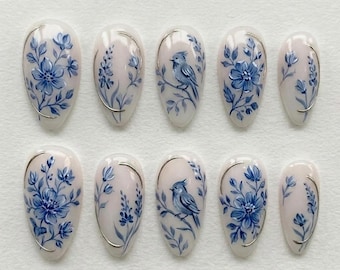 May include: Set of ten oval-shaped false nails. Each nail is painted with a light pink base and features a blue floral design, including flowers, leaves, and a bird. A silver-colored line frames each nail. The nails are arranged on a white surface.