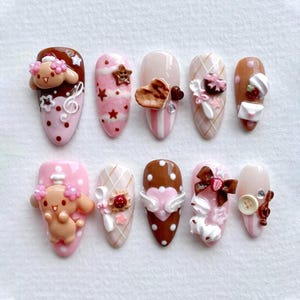 May include: A set of ten artificial nails with various designs. The nails feature a range of colors, including pink, brown, and white, and are adorned with 3D embellishments such as hearts, stars, bows, and cartoon characters. The nails are oval-shaped.
