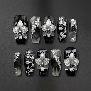 May include: A set of ten artificial nails with a black and gray marble design. Each nail features silver-colored orchid flower and leaf accents. Some nails have silver foil details, creating a glamorous look.