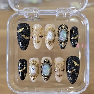 May include: A set of ten almond-shaped press-on nails in a clear plastic case. The designs feature black glitter with gold bats and stars, cream-colored nails with gold chain accents, ghost motifs in ornate gold frames, and iridescent oval gems.