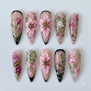 May include: A set of ten almond-shaped press-on nails featuring a nude base with intricate 3D floral designs in shades of pink, green, and gold. The nails are decorated with metallic accents, small white pearls, and black line art details.