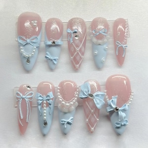 May include: A set of ten artificial nails in various designs. Each nail features a light pink base with light blue tips. Embellishments include bows, rhinestones, and pearl accents. Some nails have a glitter finish or a lattice pattern.