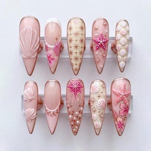 May include: A set of ten almond-shaped press-on nails featuring a coastal theme. The designs include raised 3D seashells, pink starfish, hand-painted hibiscus flowers, gold starburst patterns, and small white pearls on a soft pink and cream base.