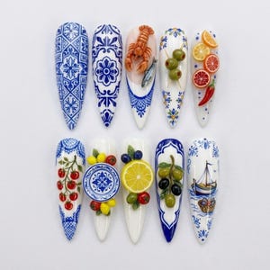 May include: A set of ten stiletto-shaped press-on nails featuring Mediterranean-inspired designs. The collection includes blue and white tile patterns, 3D miniature food motifs like lobster, olives, citrus slices, tomatoes, and a sailboat illustration.