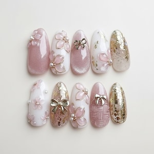 May include: A collection of ten artificial nails with various designs. Some nails are pink with glitter, others are white with pink floral accents and pearls. Gold foil and silver bows add detail. The nails are oval-shaped.