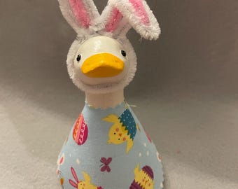 May include: A whimsical Easter decoration featuring a white duck figurine wearing a bunny ear headband with pink accents. The duck is dressed in a light blue fabric dress adorned with Easter egg and chick patterns. The duck has a yellow beak.