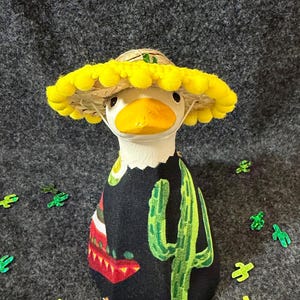 May include: A decorative duck figurine wearing a sombrero and a black poncho with a cactus design. The duck has a yellow beak and is surrounded by small green cactus-shaped decorations. The sombrero has a yellow pom-pom trim.