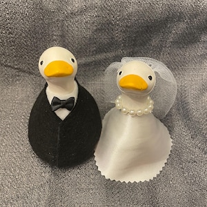 May include: Two duck figurines dressed for a wedding. One duck wears a black tuxedo with a bow tie. The other duck is dressed as a bride, wearing a white dress, pearl necklace, and a veil. The ducks have white bodies and yellow beaks.