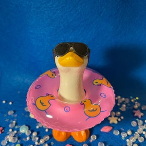May include: A whimsical ceramic duck figurine wearing sunglasses and a pink inflatable ring with duck illustrations. The duck has an orange beak and feet. The background is a solid blue color, with decorative beads and shells.