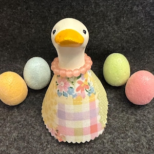 May include: A whimsical duck figurine with a white body, yellow beak, and a pink beaded necklace. It wears a patchwork dress with floral and plaid patterns. Four pastel-colored eggs surround the duck, creating a charming Easter theme.
