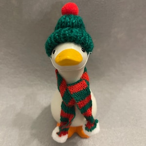 May include: A white duck figurine wearing a green knit hat with a red pom-pom, a red and green striped scarf, and a white face mask. The duck has an orange beak and feet. The figurine is a festive holiday decoration.