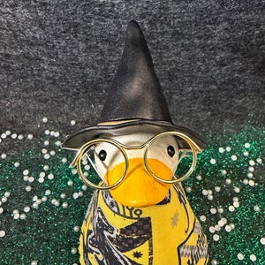 May include: A whimsical duck figurine wearing a black wizard hat and gold-rimmed glasses. The duck is adorned with a yellow and black scarf featuring a crest and the word "Hufflepuff". The background is a mix of green glitter and a dark gray surface.