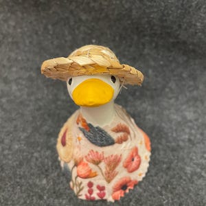 May include: A whimsical duck figurine with a straw hat and a floral-patterned dress. The duck has a white body, a yellow beak, and small black eyes. The dress features orange, red, and blue floral designs. A charming decorative item.