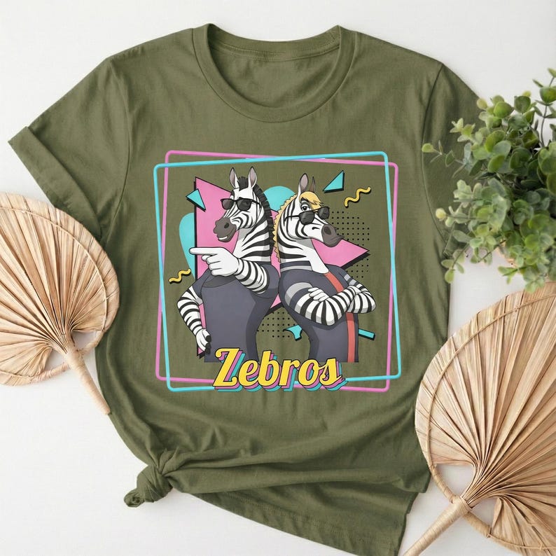 May include: Olive green t-shirt featuring a graphic of two zebras wearing sunglasses, with the word "Zebros" in yellow. The zebras are inside a pink and blue outlined box with a retro design.