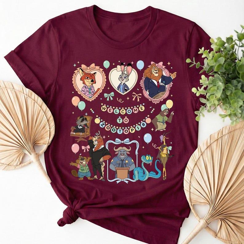 Puede incluir: Camiseta burdeos con un gr&aacute;fico colorido de personajes de Zootopia. El dise&ntilde;o incluye retratos enmarcados, globos y el texto "Welcome to Zootopia". La camiseta est&aacute; anudada en la parte inferior.